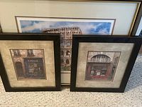 Three framed artworks displayed leaning against a wall on carpet: two smaller Paris paintings in black frames in front, one larger framed Colosseum watercolor behind them.