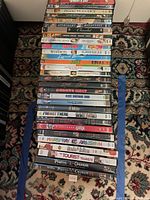Photo showing 30 DVDs stacked vertically on patterned carpet, visible titles include Dirty Dancing, Pirates of the Caribbean 1 and 2, 8 Mile, Chicago, and others.