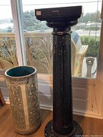 Overview of black wood pedestal and pottery umbrella stand side by side near a window, showing relative size and general appearance.
