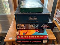 Stack of board game boxes including Trivial Pursuit, Cranium, Battleship, and magnetic playing cards on a stool.