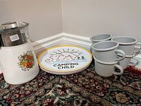 Photo showing vintage Corningware coffee percolator, camping plates, and enamel cups together on carpeted floor.