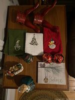 Photo of Christmas-themed tea towels, red baubles, snow globe, decorative sign, and cast iron tree stand on wooden table.