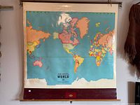 Full view of the vintage world map hanging on a wall showing colorful country outlines on blue ocean background, mounted on wooden rollers at top and bottom.