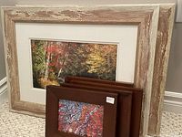 Four small dark brown wood framed prints with fall tree scenes stacked in front of two large white rustic wood framed fall landscape prints on carpeted floor.