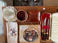 Overview of all items on wooden table: vintage teacup and saucer, three small wooden bowls (one dark, one mid-brown, one reddish), small square wooden dish, Norman Rockwell numbered plate in plastic sleeve, embroidered table runner folded under plate, carving set case closed with fabric box.