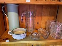 Overall view of shelf containing ceramic thermos pitcher, clear glass pitcher, ceramic handled bowl, crystal shallow bowl, rectangular crystal coaster, and two clear glass vases