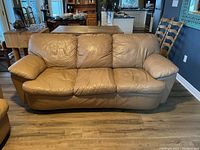 Front view of a tan leather sofa with three cushions and padded armrests, situated on wood flooring with some surrounding furniture visible.