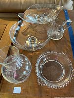 Overview of crystal pieces including gravy server, divided bowl, vase, raised fruit bowl, and candy bowl on a wooden surface for condition and completeness assessment.