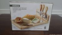 Box showing product front with image of the complete cheese platter set including cheese board, magnetic wooden knife holder, and four cheese knives.