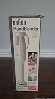 Photo showing the full front of the boxed Braun Hand Blender, white color with handle and control window visible.