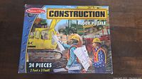 Box top showing illustration of construction floor puzzle with two workers holding blueprints and heavy machinery in the background.
