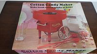 Boxed red cotton candy maker machine with clear plastic cones and red scoops visible in the packaging image.