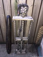 Front view showing the carved African figure CD holder next to the dark carved Indonesian tribal mask, both leaning against a wooden fence outside.