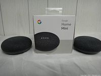 Two charcoal Google Home Mini smart speakers flanking one retail box showing product image and Google branding.