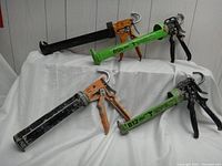 Four cartridge caulking tools: two large with orange handles and black/orange frames, and two small with black handles and green frames labeled B12.