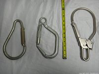 Three large metal snap hooks and carabiner side by side with measuring tape to show size, approximately 10-11 inches each.