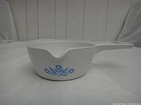 Side view of white Corning Ware saucepan with blue cornflower pattern, showing handle and pouring spout, set on plain surface with white backdrop.