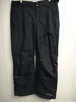 Front view of black Columbia ski pants hanging, showing full length with belt loops, pockets, and logo near hem.