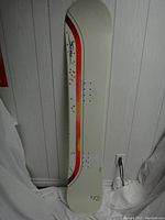 Side view of snowboard standing vertically against wall, showing mounting holes and Sims logo.