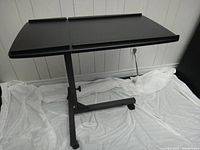 Black adjustable over bed table with tilting top surface and height adjustment, shown from front angle.