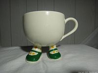 Front view of the vintage ceramic cup showing the attached feet with green shoes and yellow polka dot socks.