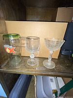 Two handcrafted glasses displayed beside an empty beer glass with a green rim on a wooden shelf