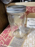 Single clear glass jar with metal lid, contoured shape, sitting on cardboard box.