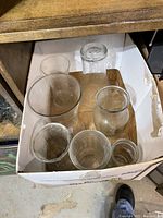 Box with seven clear glass vases of various shapes and sizes inside cardboard box on floor.