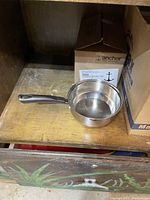 Stainless steel saucepan with black handle on wooden shelf next to a box labeled Anchor