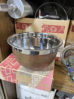 Large metal mixing bowl sitting on patterned box, showing shiny metal surface with some scratches and marks.