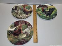 Three round decorative plates showing different rooster and chicken farm scenes with ruler for scale.