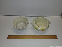 Two ceramic flower pots with ruler for scale, white pot with scalloped edge and round pot with yellow flower motif.