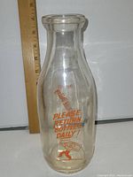 Front view of a tall clear glass milk bottle next to a ruler showing a height of approximately 12 inches. Bottle has orange printed text "PLEASE RETURN BOTTLES DAILY!" and a running milkman graphic.