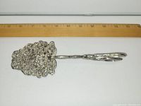 Side view of the antique silver plated cast metal pie lifter placed adjacent to a ruler showing approximately 9 inches in length. The lifter head has intricate ornate openwork design.