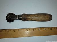 Top view of the antique roller tool showing wooden handle and spiked metal roller with ruler for size comparison at about 8 inches.