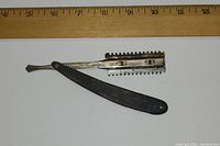 Photo showing antique straight razor with black handle and metal comb safety guard, laid flat against a white background with a wooden ruler above it indicating approx. 5 inches length.