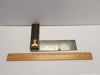 Antique woodworking square laid horizontally with a wooden ruler underneath to show measurement; features a hardwood handle with brass inlay and a metal blade.