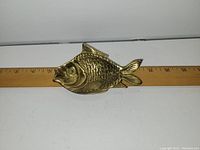 Top down view of small brass fish-shaped bowl with textured scales and fins, placed next to a ruler for scale, showing length about 4.75 inches.