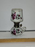 Front view of small ceramic oil lamp with floral design and matching shade, next to ruler for size reference.