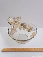 Two clear glass bowls, larger bowl and smaller bowl for dip, decorated with gold leaves and berries and mounted on a black metal stand, placed beside a wooden ruler for scale.
