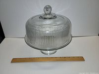 Vintage clear thick glass cake stand with vertical ribbed dome lid and round knob handle, shown with a ruler for scale approximately 12 inches diameter and 10 inches tall.