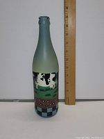 Front view of a 12-inch tall hand painted glass bottle showing a black and white cow in a farm scene against a muted green and blue background with checkered pattern at the bottom.