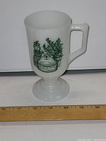 White milk glass mug with green spice jar and herb graphic on front side, placed beside a ruler showing 6 inches height.
