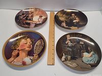Four Norman Rockwell artist collector plates arranged around an 8 inch ruler to show size. Plates feature classic Rockwell scenes with people in nostalgic settings.