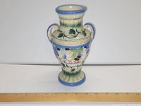 View of vase with decorative handles, blue rim and foot, a painted classical relief scene, and green drip glaze on cream body.