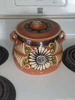 Front view of the terracotta bean pot sitting on a stovetop, showing detailed floral and spiral painted design and lid with knob.