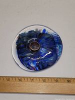 Top view of circular blown glass candle holder showing blue swirl pattern and clear glass with a copper pipe center. A ruler below shows it measures about 5 inches in diameter.