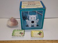 Photo showing two small square ceramic bowls, one pale green and one pale yellow with seashell and rose design, alongside the original boxed ceramic cat figurine on swing.