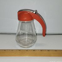 Front view of vintage glass maple syrup jug sitting on white surface with ruler in front showing approximately 5.5 inches tall. Red handle and metal sliding lid clearly visible.