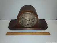 Front view of the wooden mantel clock with a curved top, round clock face with worn dial, no glass cover, and ruler for size reference.